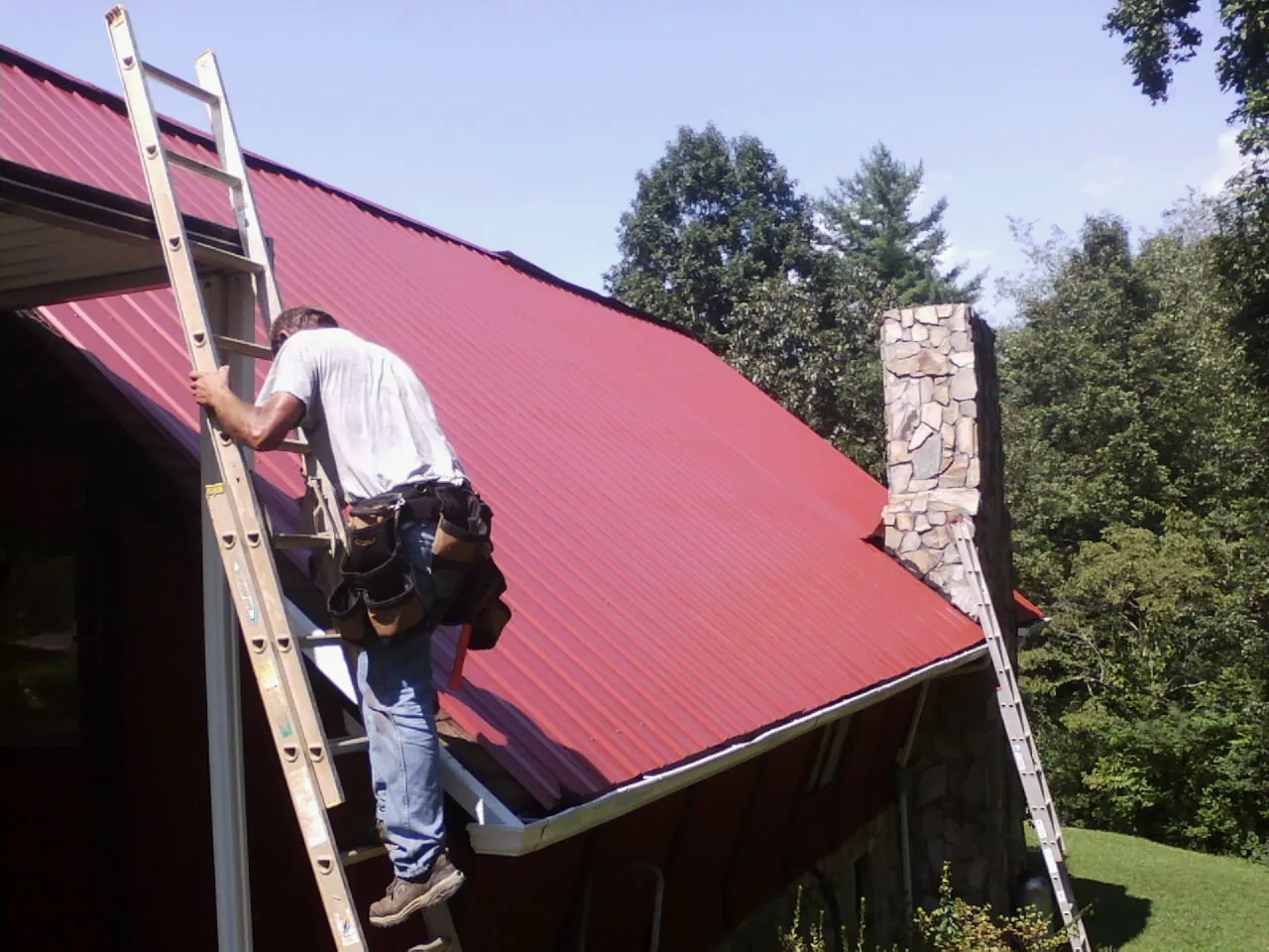 Roofer on a ladder beside a red metal roof and stone chimney — Soffit & Fascia Repair by Roofing Brothers in Pilot Mound